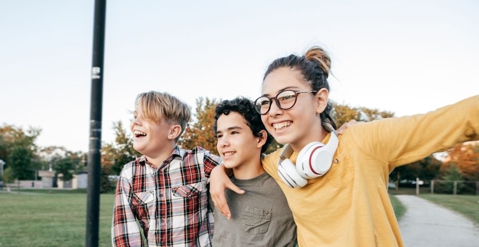 Three smiling kids standing together outdoors, with one wearing glasses and headphones, enjoying a sunny day at the park